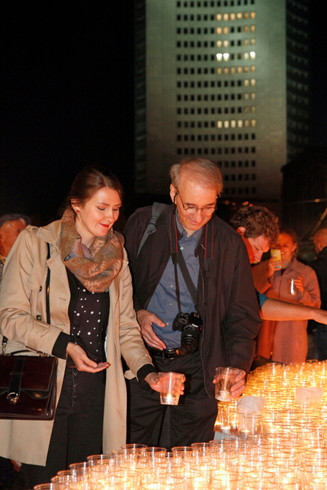 Ein Mann und eine Frau stellen jeweils ein Glas mit einer Kerze darin auf ein Podest, auf dem bereits viele leuchtende Gläser stehen.