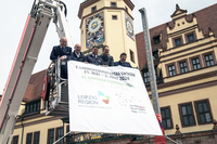 Vier Männer auf einer Drehleiter der Feuerwehr halten ein Banner mit der Ankündung zum 7. Landesfeuerwehrtag Sachsen hoch.