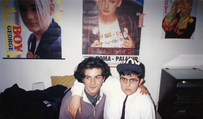 Zwei queere Teenager im Jahre 1988 auf der Couch sitzend mit Boy George-Postern im Hintergrund.