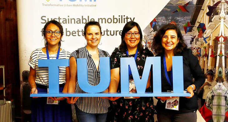 Vier Frauen halten einen großen Blauen Schriftzug "TUMI" hoch und lächeln in die Kamera.