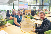 Zwei Frauen und ein Mann drehen sich lächelnd von ihren Sitzen in einem großen Plenarsaal ins Bild. Vor Ihnen steht eine runde, goldene Skulptur.