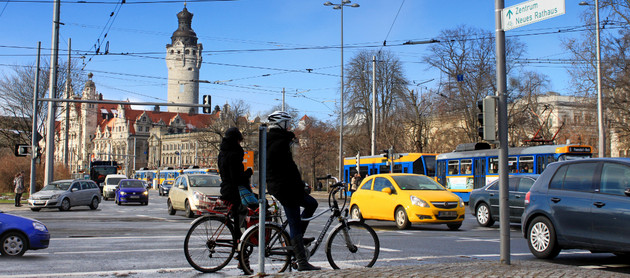 Blick auf eine große Straßenkreuzung in Leipzig mit Autos, Radfahrern und Straßenbahnen. Im Hintergrund das Neue Rathaus.
