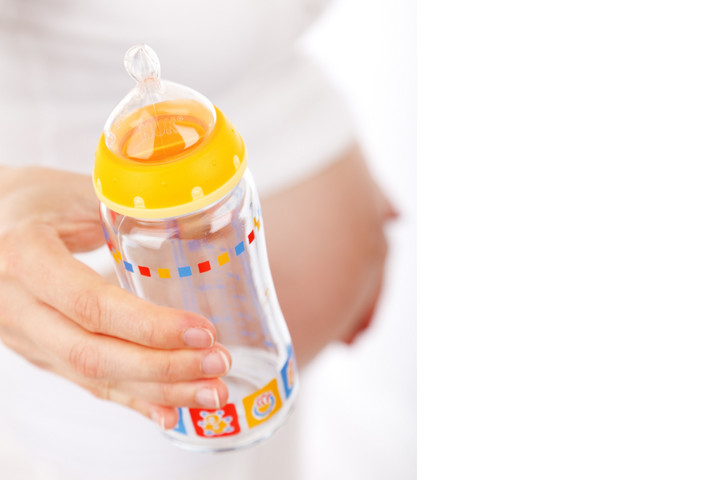 Hand hält eine Babyflasche. Im Hintergrund ist ein Babybauch
