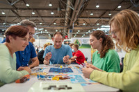 Fünf Menschen sitzen um einen Tisch in einer Messehalle und spielen ein Gesellschaftsspiel