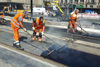 Bauarbeiter in orangefarbener Schutzkleidung bearbeiten mit Werkzeugen den Asphalt zwischen zwei Gleisen.