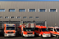 Vor einem rechteckigen Feuerwehrgebäude stehen vier Feuerwehrfahrzeuge und ein Krankentransportwagen.