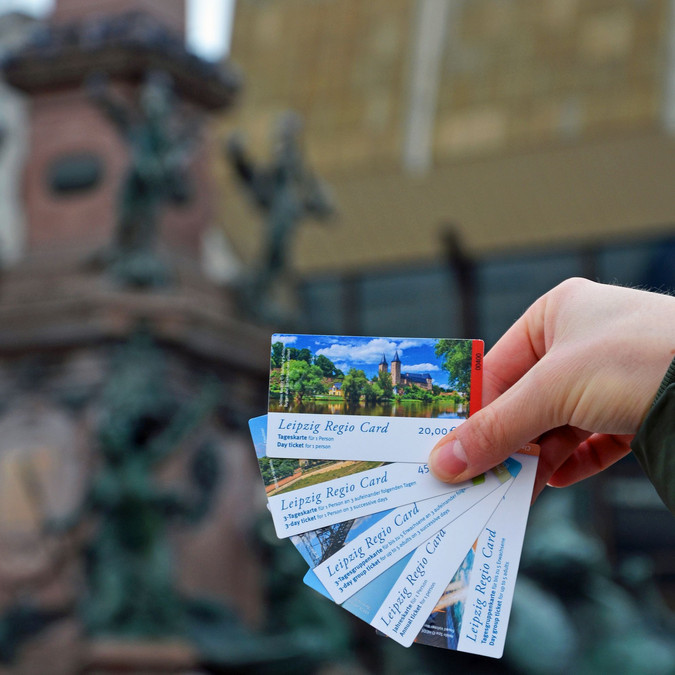 Eine Hand hält fünf verschieden Regio Cards. Im Hintergrund ist der Mendebrunnen auf dem Augustusplatz.