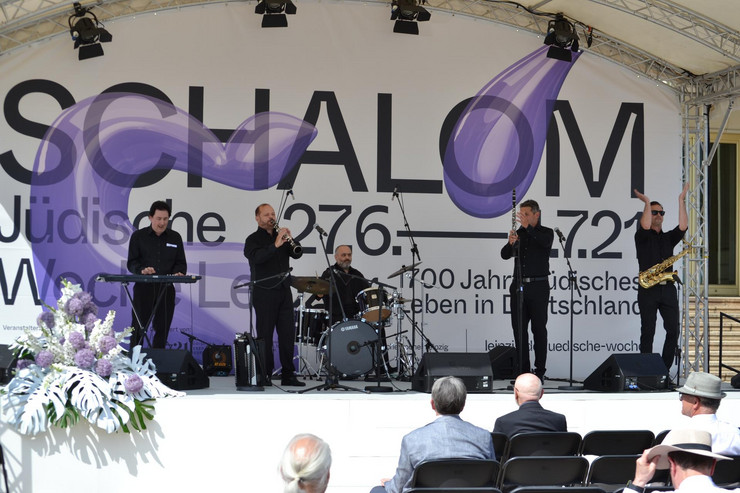 Fünf Musiker stehen auf einer großen Bühne und spielen Klarinette, Keyboard, Schlagzeug, Saxophon und singen.