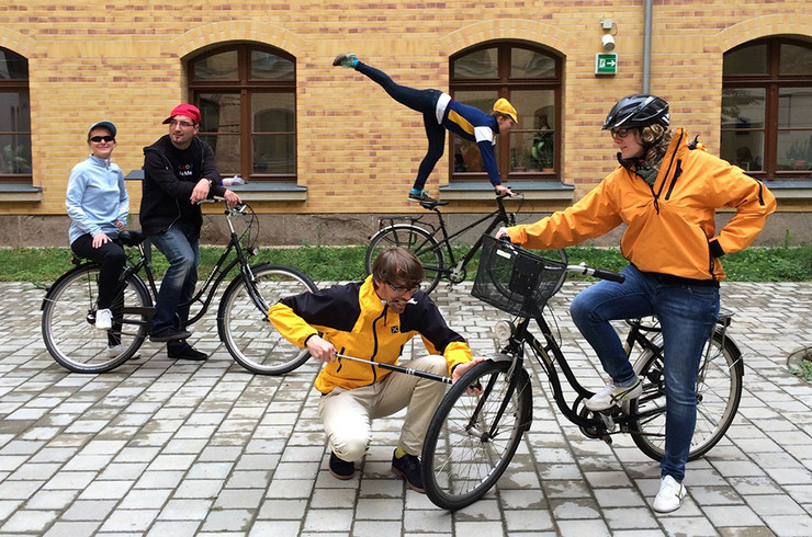 Foto: Hof der Stadtbibliothek vor der Fassade, links Mann auf Fahrrad, Frau auf Gepäckträger, rechts Frau auf Fahrrad, Mann pumpt Luft auf Vorderrad, im Hintergrund rollt Frau artistisch stehend auf Sattel eines Fahrrades