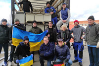 Elf Männer und zwei Frauen stehen vor der Ladefläche eines Lkw. Sie halten zwei ukrainische Flaggen und eine Deutsche in den Händen. Auf der Ladefläche sind zahlreiche Krankenhausbetten und Nachttische verstaut. 