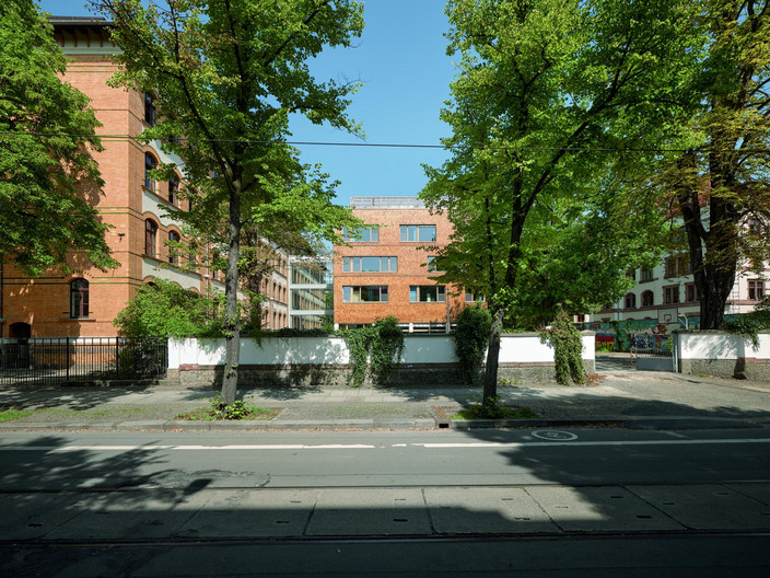 Blick von der Straße auf die Schule, den gläsernen Verbindungsgang und den Erweiterungsbau, teilweise durch im Vordergrund stehende Bäume verdeckt.
