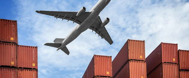 Frachtcontainer, darüber fliegt ein großes Flugzeug vor blauem Himmel