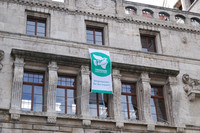 Eine grün-weiße Flagge mit der Aufschrift "Bürgermeister für den Frieden" hängt an einem steinernen Gebäude mit mehreren Fenstern. Eine weiße Taube vor einem grünen Kreis ist auch darauf abgebildet. 
