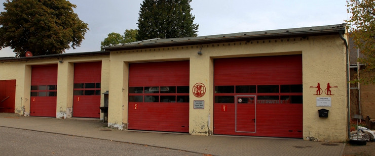 Vier gelb angestrichene Garagen mit roten Rolltoren. An den Wänden sind zwei rote Feuerwehrsymbole und ein Briefkasten. Im Hintergrund stehen Bäume.
