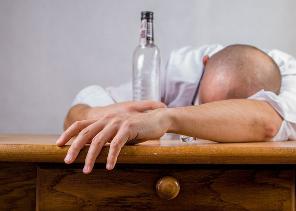 Ein Mann liegt mit dem Gesicht nach unten auf einem Tisch. In der einen Hand hält er eine leere Flasche, die andere ist ausgestreckt.