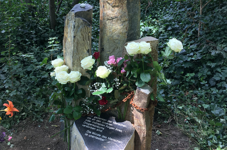 Zwei steinerne Stelen mit Blumen geschmückt. Davor eine Tafel mit der Inschrift "Wir gedenken der Menschen, die an den direkten oder indirekten Folgen ihres Drogenkonsums verstorben sind. Die Würde des Menschen ist unantastbar"