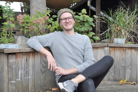 Ein lächelnder junger Mann mit Brille und Mütze im grauen Pulli auf einer Holzbank