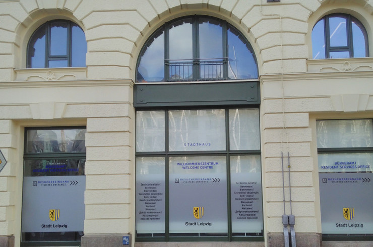 Das Erdgeschoss eines hellgelben Gebäudes mit großen Fenstern und einer gläsernen Eingangstür, auf denen das Logo der Stadt Leipzig zu sehen ist