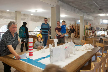 Kinder und Erwachsene stehen vor einer aus Papier und anderem Material gestalteten Stadt, die auf einem großen Holztisch steht.