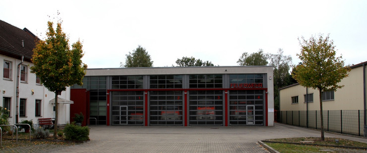 Eine große Halle mit vier Toren aus Glassegmenten. In der Halle stehen vier Feuerwehrfahrzeuge und über dem rechten Tor steht in großen roten Buchstaben das Wort "Feuerwehr".