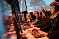Mehrere Computerspieler sitzen in einer Reihe nebeneinander an ihren Rechner und halten ihre Hände auf den Tastaturen. Die meisten haben Kopfhörer auf und schauen auf ihre Monitore.