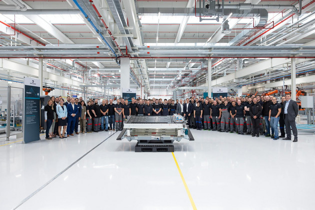 Blick in die Produktionshalle des Dräxlmaier-Werks in Leipzig mit vielen Mitarbeitern, die im großen Halbkreis um ein Bauteil stehen.