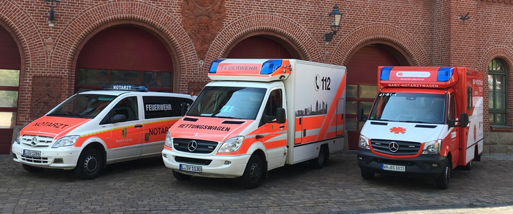 Drei Rettungsdienstfahrzeuge stehen nebeneinander, ein Notarzteinsatzfahrzeug, ein Rettungstransportwagen und ein Baby-Notarztwagen. Im Hintergrund ist ein Backsteingebäude mit bogenförmigen Toren.