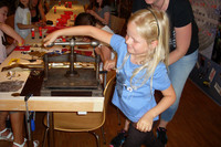 Ein blondes Mädchen steht an einer großen Metallpresse. Im Hintergrund sieht man Kinder an einem langen Tisch mit allerlei Bastelmaterialien.