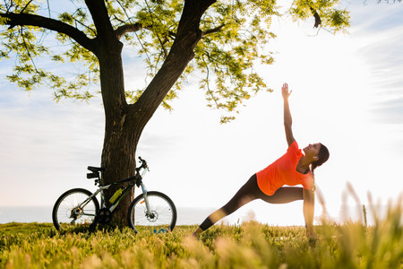 Junge Frau, die im Freien Yoga-Übungen macht, ihr Fahrrad in an einem Baum gelehnt