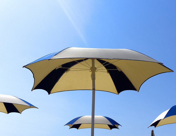blau-weißer Sonnenschirm vor einem blauen Sommerhimmel. Im Hintergrund sind drei gleiche Sonnenschirme