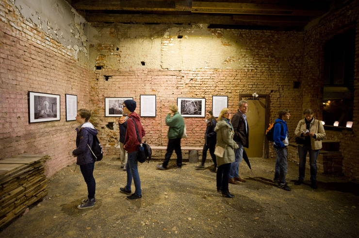 Besucher schauen sich Fotografien und Kunstwerke an, die an den Wänden hängen. Der Raum verbreitet einen industriellen Charme durch die sichtbaren Ziegelsteine der Wände.
