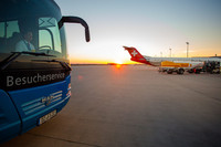 Ein Bus mit der Aufschrift Besucherservice steht auf dem Flughafengelände. Daneben steht ein Flugzeug mit einem Tankwagen. Am Ende des Geländes geht die Sonne unter.