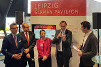 Oberbürgermeister Jung, umringt von weiteren Personen, spricht in ein Mikro. Die Gruppe steht vor einem großen Aufsteller, auf dem in Großbuchstaben "Leipzig, German Pavilion" steht.