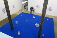 Ausstellungsraum mit einer großen blauen Fläche, auf der mehrere kleine Ausstellungsobjekte in Glaswürfeln stehen. Zwei Besucher schauen sich diese an.