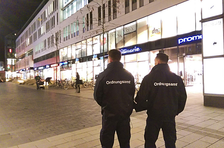 Zwei Männer in dunkler Uniform mit der Aufschrift "Ordnungsamt", von hinten fotografiert mit Blick in eine Fußgängerzone