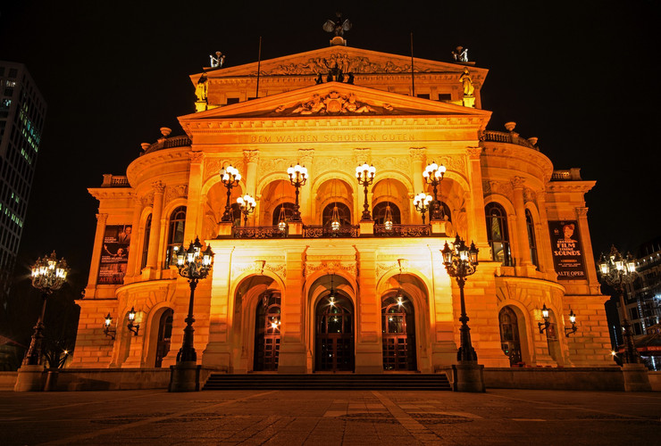 Frontansicht der Alten Oper mit drei großen Eingangsbögen und einer Terasse darüber. Am Giebel steht geschrieben "Dem Wahren Schönen Guten".