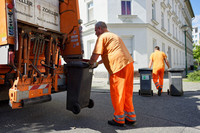 Ein Mitarbeiter der Stadtreinigung hängt eine Mülltonne zur Entleerung an ein Müllauto. Ein weitere Mitarbeiter bringt zwei Mülltonnen zum Abstellplatz.