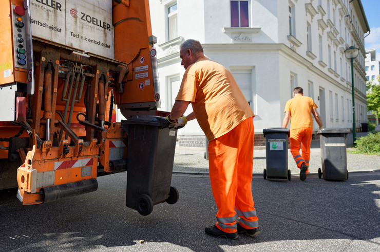 Ein Mitarbeiter der Stadtreinigung hängt eine Mülltonne zur Entleerung an ein Müllauto. Ein weitere Mitarbeiter bringt zwei Mülltonnen zum Abstellplatz.