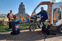 Mehrere Menschen mit Müllbeuteln, eine Mülltonne und ein Lastenrad auf einer Grünfläche vor einem Denkmal