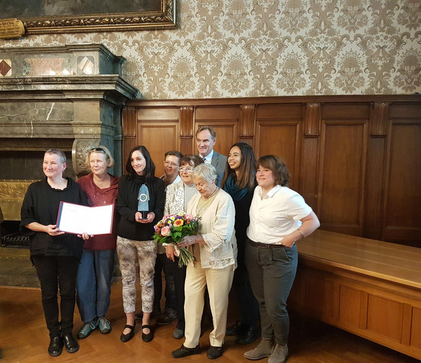 Acht Frauen der Frauenkultur Leipzig und der Oberbürgermeister stehen in einer kleinen Gruppe zusammen und halten in den Händen die Urkunde, den Preisgegenstand und einen Blumenstrauß.
