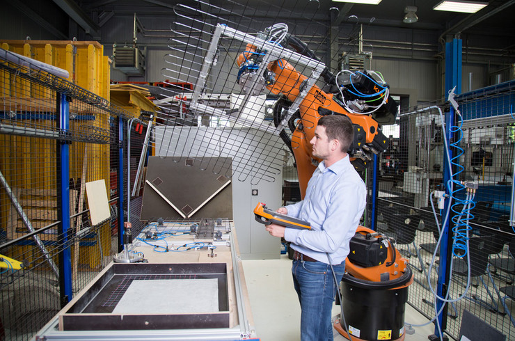 Ein Mann steht in einer Werkstatthalle voller technischer Geräte und bedient einen Roboterarm, der ein Drahtgeflecht bewegt.