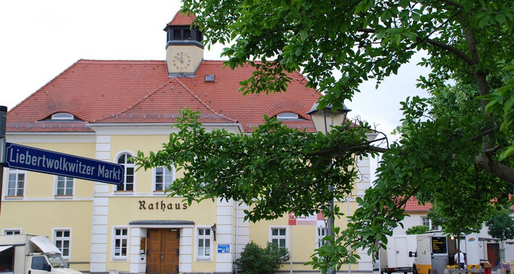 Ein weiß-gelbes Gebäude mit Türmchen und Uhr. In alten Lettern steht über dem Eingang "Rathaus". Daneben werden Marktstände aufgebaut.