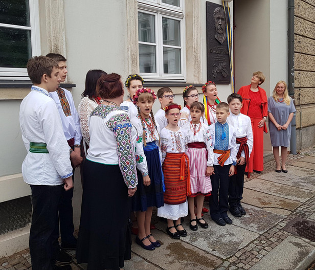 Vor einer grauen Hauswand steht ein Kinderchor in traditioneller ukrainischer Kleidung und singt. Die Mädchen mit bunten Blumenkränzen auf dem Kopf und Schleifen im Haar. An der Hauswand ist eine bronzene Gedenkplatte zu sehen.