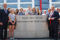 Viele Mädchen und einige Erwachsene stehen hinter beziehungsweise neben einem großen Eingangsschild aus Metall, auf dem "Gerda-Taro-Schule - Gymnasium der Stadt Leipzig" steht. Drei der Mädchen halten historische Fotoapparate in der Hand.