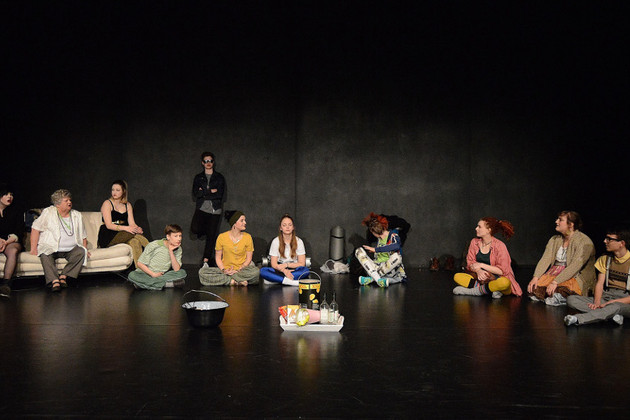 Elf Darsteller sitzen mit Getränken und Chipspackung im Halbkreis auf der Bühne bzw. auf einem Sofa