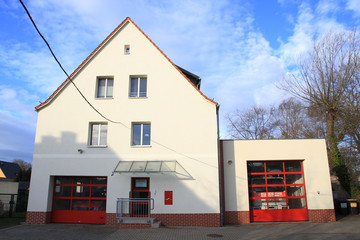 Ein weißes Haus mit großem Spitzdach. Unten links ein rotes Rolltor und in der Mitte eine Eingangstür. Darüber zwei Etagen mit jeweils zwei Fenstern. Auf der rechten Seite ein flacher Anbau mit einem großen, roten Rolltor mit Fenstern.