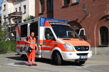 Rettungswagen der Berufsfeuerwehr. Davor steht ein Rettungsdienstmitarbeiter in oranger Arbeitskleidung mit einem medizinischen Gerät über der Schulter.