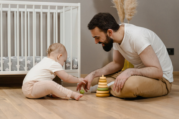 Ein Mann und ein Baby sitzen sich auf dem Boden gegenüber. zwichen den beiden ist Holzpielzeug. Im Hintergrund ein Gitterbett.