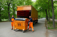 Zwei Mitarbeiter der Stadtreinigung stellen eine große orangene Mülltonnen auf. Dahinter steht ihr Transportfahrzeug.