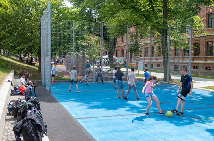 Kleines Fußballfeld mit Minitoren auf denen mehrere Kinder spielen. Im Hintergrund ein Gymnasium und viele Bäume.